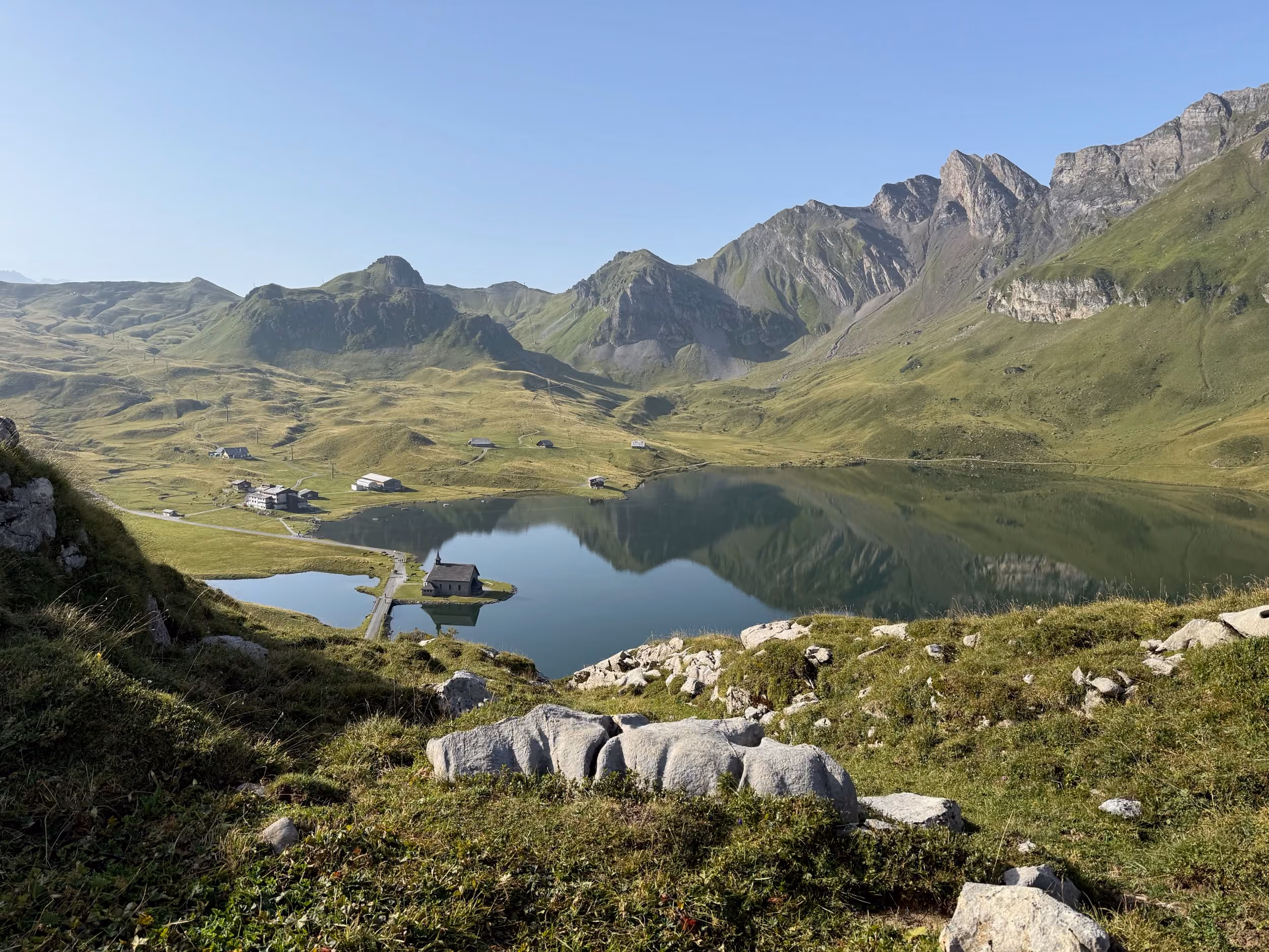 Blick vom Bonistock auf den Melchsee mit der Kapelle, dahinter grüne Alpweiden und Gipfel